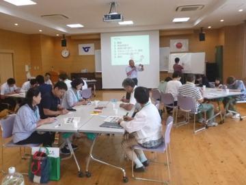 スクリーンの前で話されている方がいて、出席した委員の方々が席に着いて聞いている写真