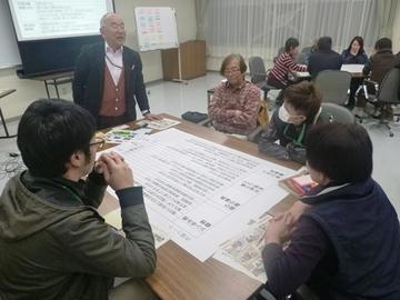 資料がテーブルに置かれてあり、年配の男性が立って話をしているのを聞いている4人の男性の写真