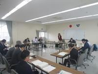 有識者懇談会で奥のテーブルの女性が立ち上がっていて他の参加者の人達は資料を見ている写真
