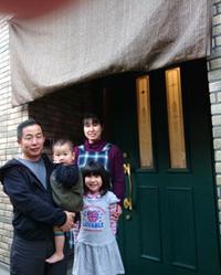緑色の玄関扉の前で小さな赤ちゃんを抱いた宮崎さんとその横に並ぶ娘さんと奥さんの家族写真