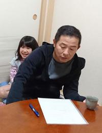 テーブルに紙とペン、湯飲みを置き考えている宮崎さんの後ろでニコニコ顔の娘さんの写真