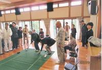 1人の高齢者の方が緑のマットの上で遊技をするのを周りで見ている高齢者の方々の写真