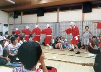 畳の中央で踊っている赤い衣装を着たひょっとこを見ている参加者達の写真