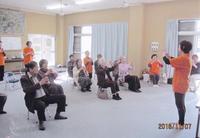 オレンジのお揃いのTシャツをきた人達が一人暮らしの高齢者の集いで体操を教えている写真
