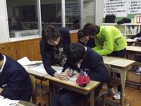 各机に座る生徒一人一人に指導者の人達が横に立ちながら勉強を教えている写真