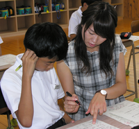 1人の女子学生がペンを持ちプリントを指さし男子生徒に教えている写真