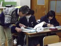 女子学生2名が並んで机で勉強しているところを男性の指導者がノートをのぞき込んで教えている写真