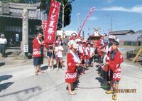 赤生地に白文字でよど夏祭りと書かれた旗を持つ人の前にたくさんの人達がお揃いのハッピを着て2列に向き合って並び神輿が来るのを待っている写真