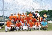 グラウンドでオレンジと白のユニフォーム姿の壮年ソフトボール部の人達を写した集合写真